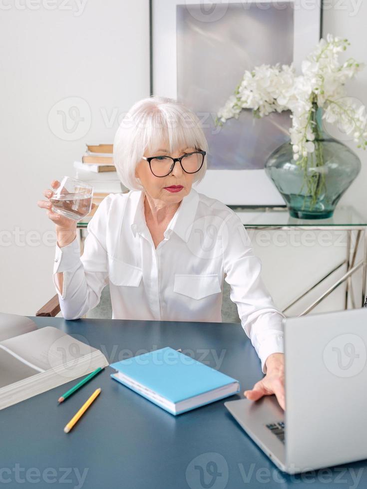 Mujer de cabello gris hermosa senior en blusa blanca bebiendo agua durante el trabajo en la oficina. trabajo, personas mayores, equilibrio hídrico, encontrar una solución, concepto de experiencia foto