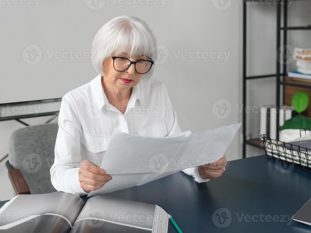 Mujer de cabello gris hermosa senior cansada en blusa blanca leyendo documentos en la oficina. trabajo, personas mayores, problemas, encontrar una solución, concepto de experiencia foto