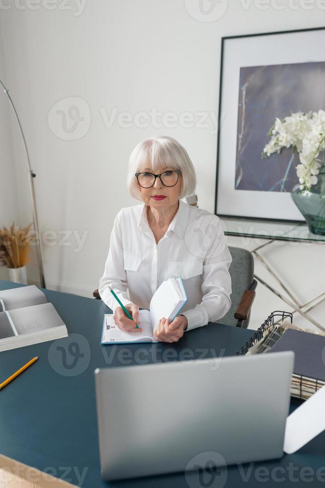 Mujer de cabello gris hermosa senior cansada en blusa blanca leyendo documentos en la oficina. trabajo, personas mayores, problemas, encontrar una solución, concepto de experiencia foto