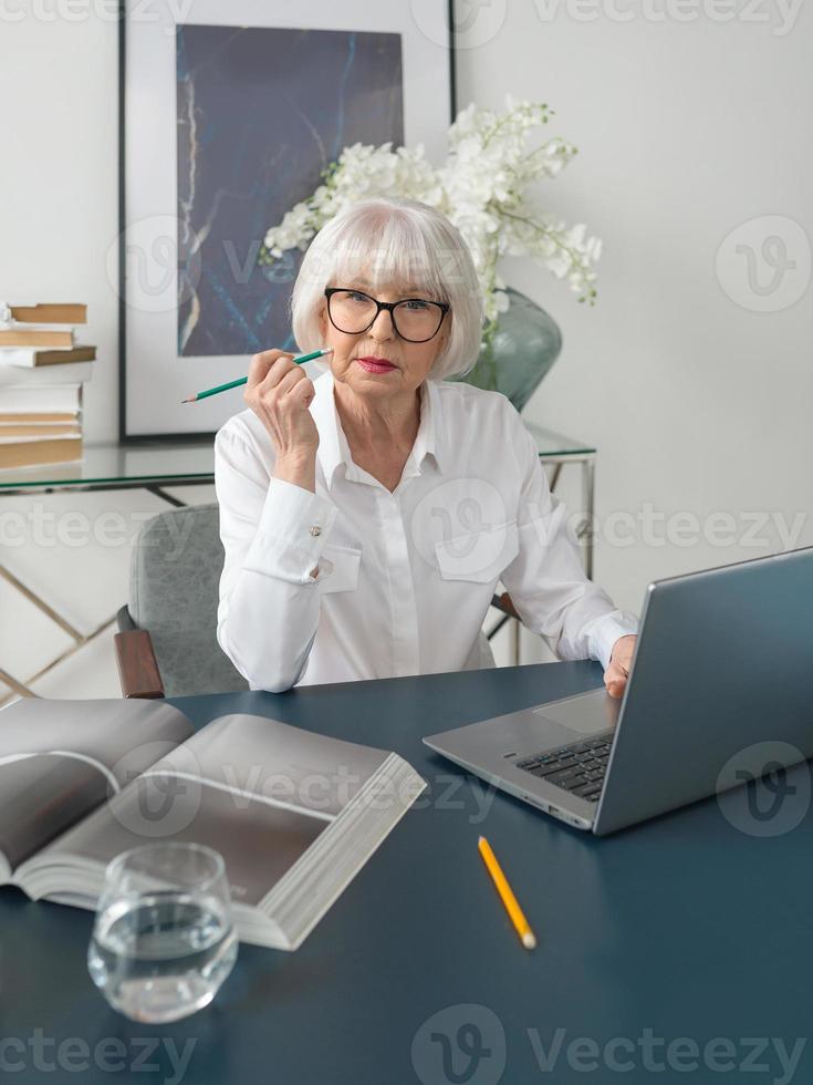 Mujer de cabello gris hermosa senior cansada en blusa blanca leyendo documentos en la oficina. trabajo, personas mayores, problemas, encontrar una solución, concepto de experiencia foto