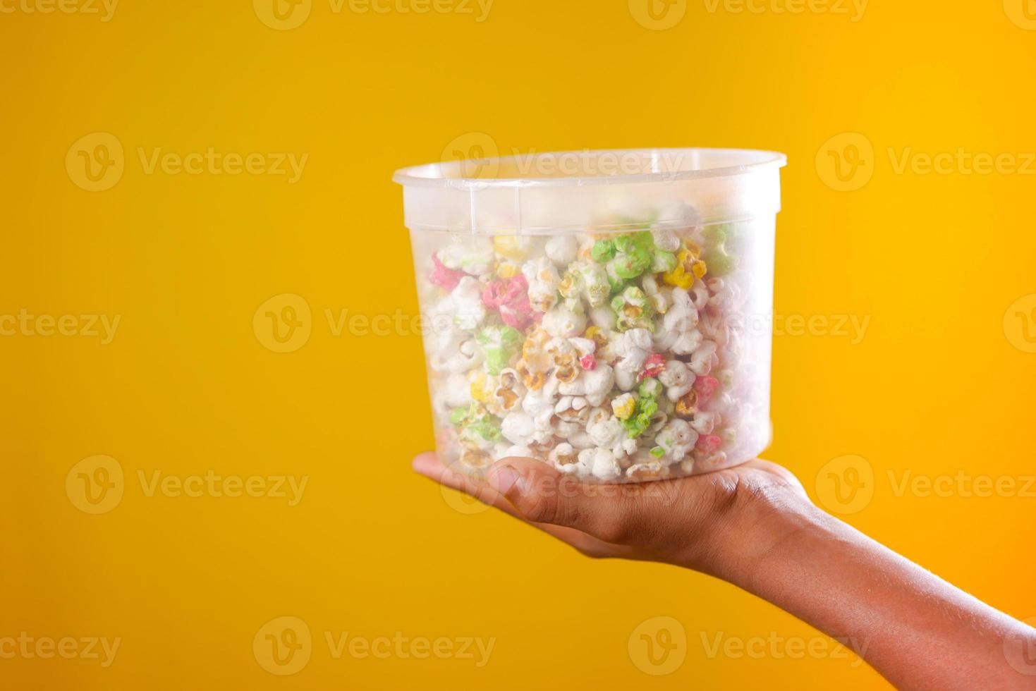 Asimiento de la mano un recipiente de palomitas de maíz contra el fondo amarillo foto