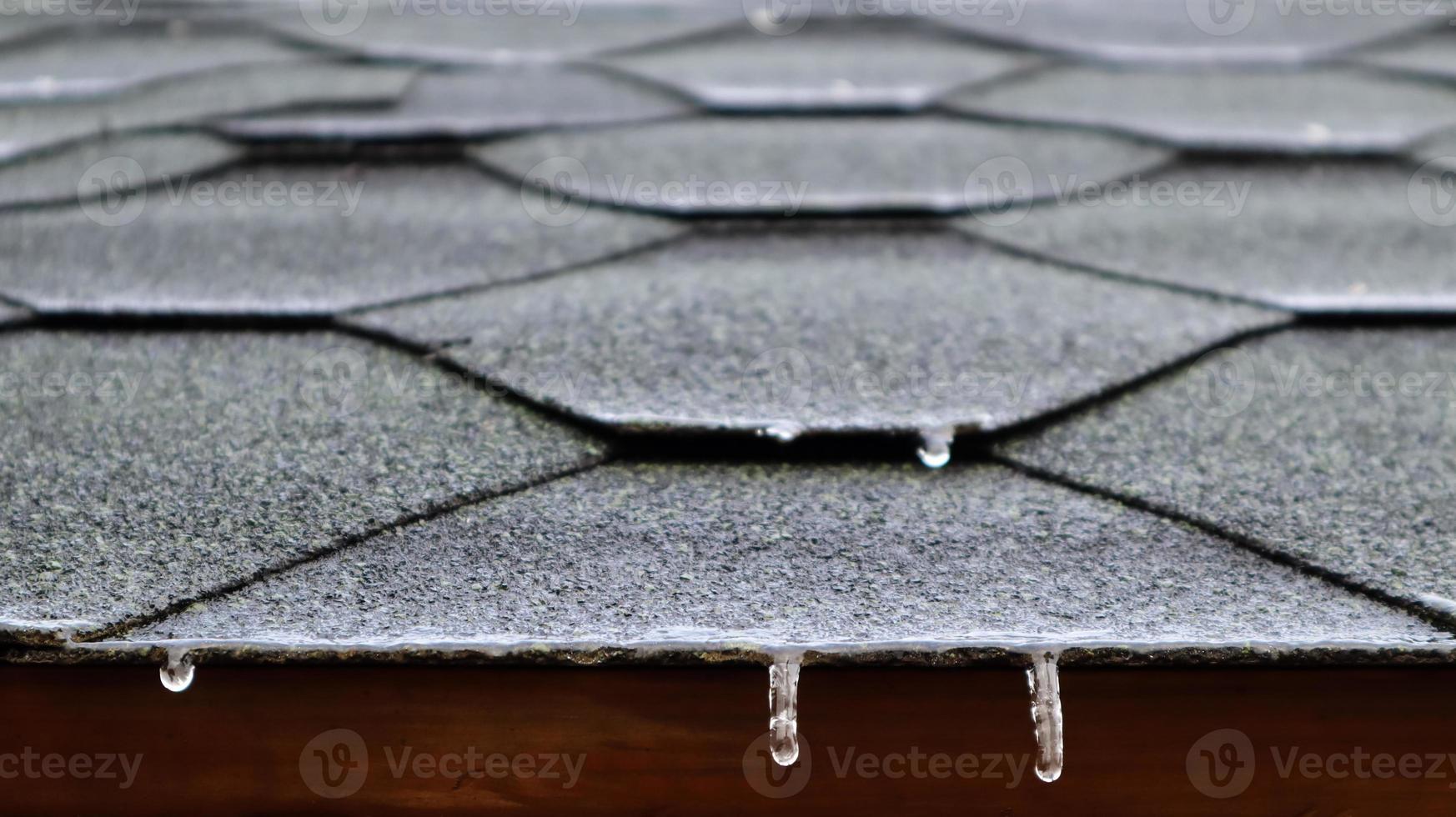Roofing on the roof of a house or gazebo made of bituminous tiles with frozen flowing water and hanging icicles. Close-up. photo