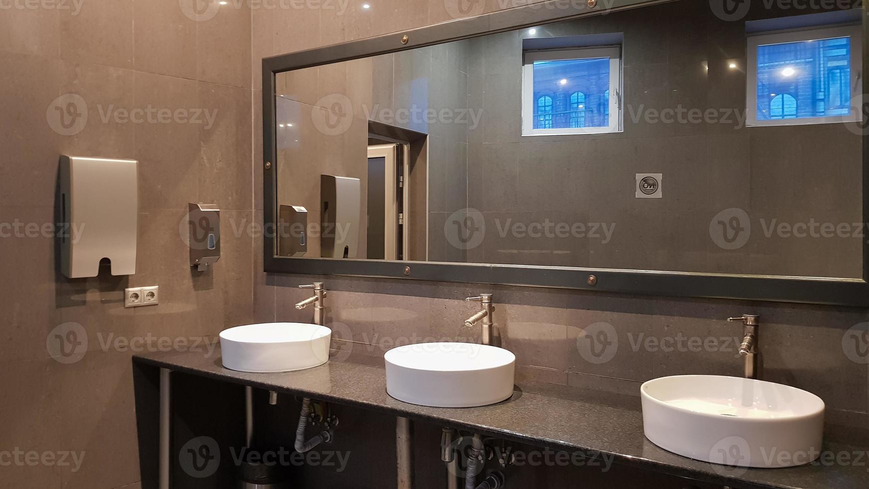 Chrome faucets with white round washbasins in a public toilet with a large mirror and gray walls, modern interior of a public toilet. photo