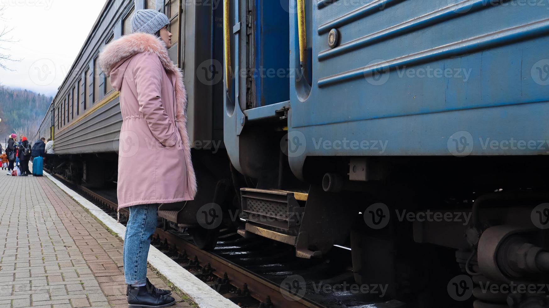 A woman in a gray knitted hat and a pink jacket with fur enters the carriage of an old train at the station. Rail transportation. Autumn and winter travel concept. Railway transport. photo