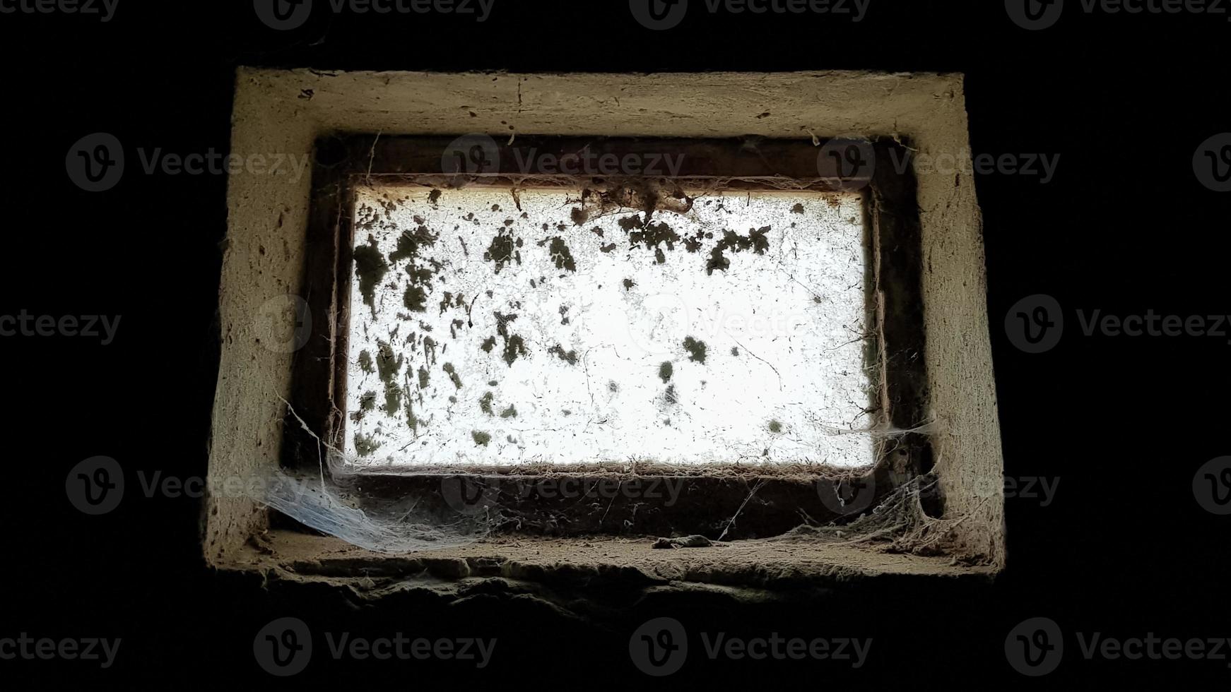 a small rectangular dirty and dusty web window in a large basement. White light penetrates the dark basement. photo