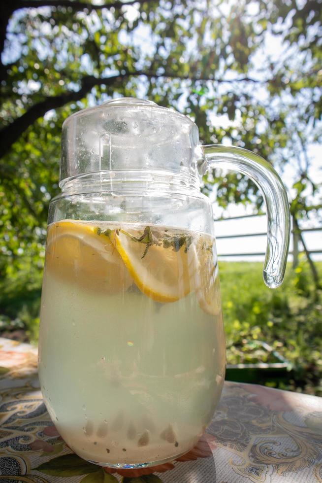 Homemade lemonade made from lemons in a large glass jug on the table in the garden. A jug with lemon and mint stands on the street against the backdrop of greenery on a hot summer day. photo