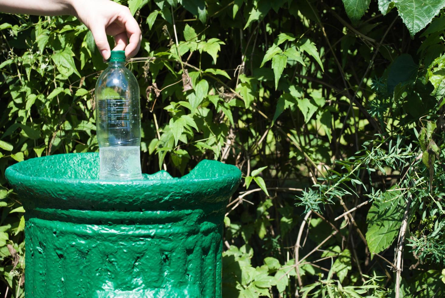 Female hand throws a plastic bottle into a small iron trash can in a green park in summer, close up. nature protection from plastic. photo