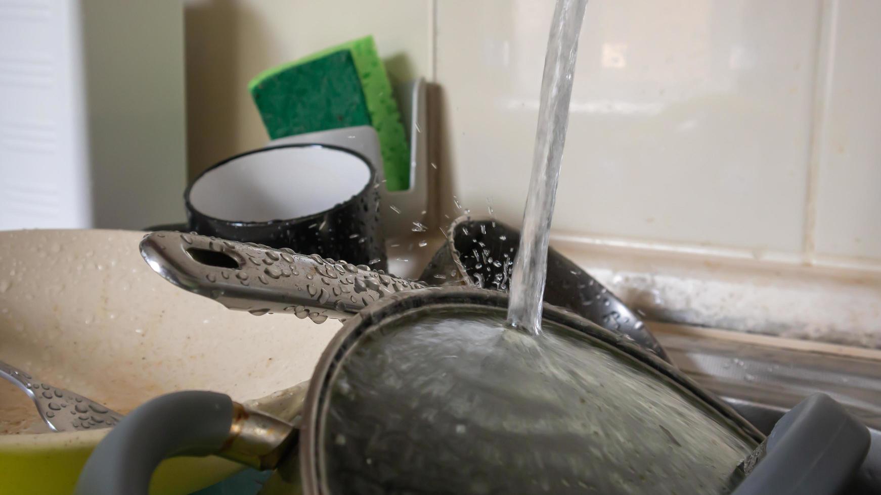 The kitchen utensils in the wash basin need to be washed. A pile of dirty dishes in the kitchen sink with running water. Kitchen utensils need washing. Homework concept. photo