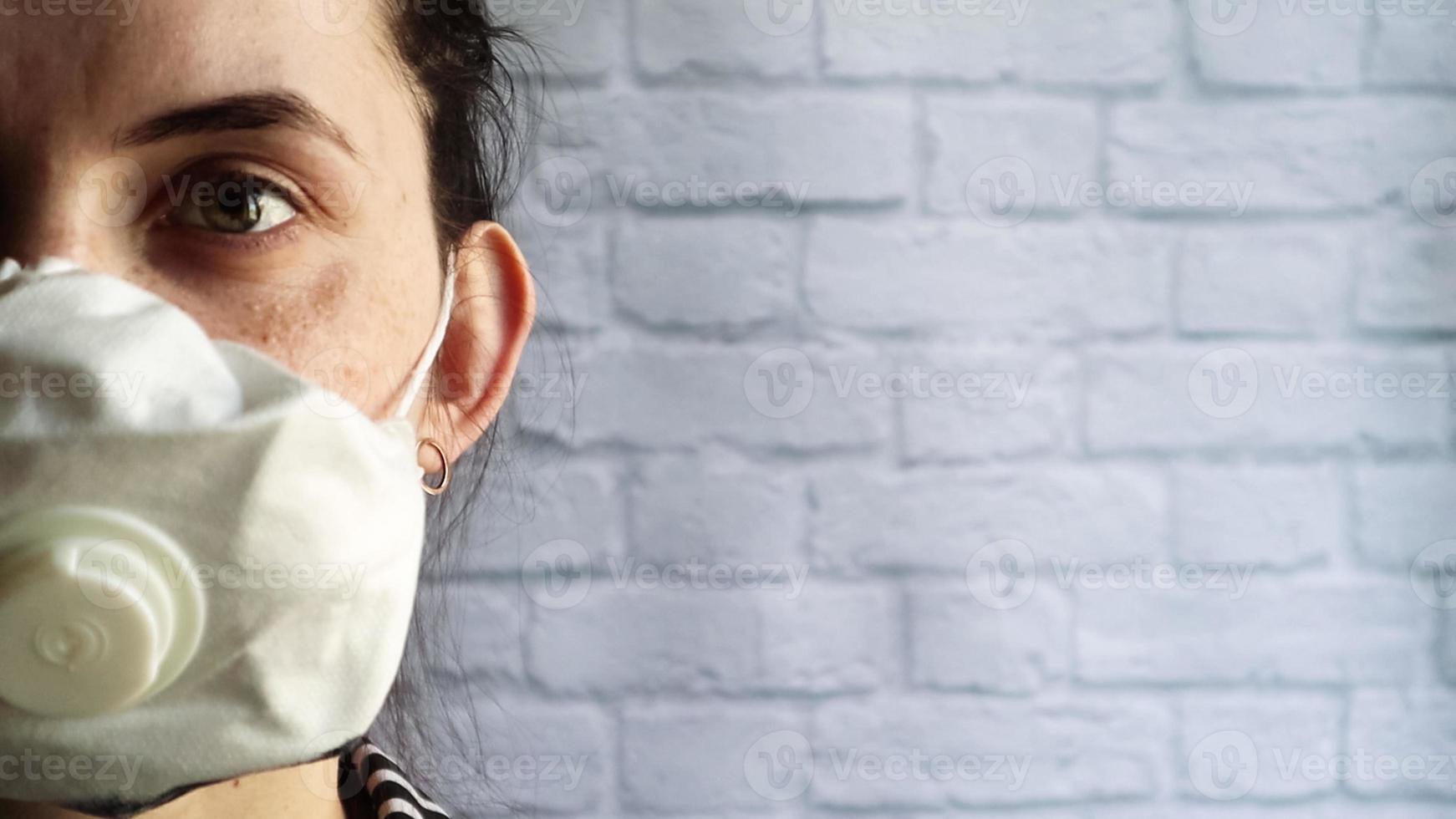 Close up half face of an upset caucasian young woman in medical mask, looking directly at the camera, standing against a white brick wall with copy space. Air pollution concept. coronavirus concept. photo