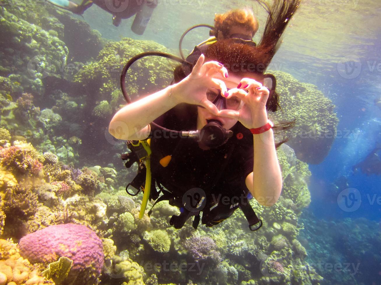 La encantadora niña buceadora muestra con un gesto de la mano un signo de amor, un corazón de los dedos con un instructor asociado para nadar de forma segura bajo el agua en el mar rojo con arrecifes de coral Egipto, Sharm el Sheikh foto