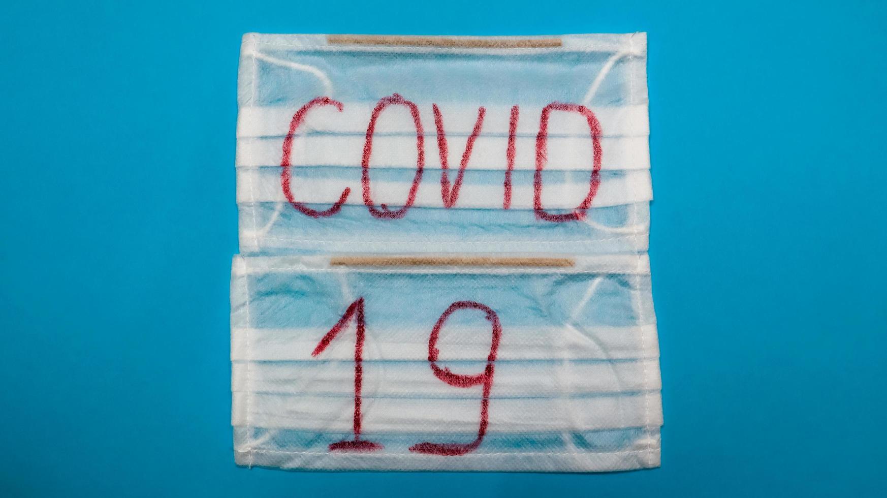 Máscara quirúrgica blanca típica de 3 capas con ganchos de goma para cubrir la boca y la nariz con letras mayúsculas en inglés covid-19 sobre un fondo azul. virus peligroso y concepto de cuarentena. foto