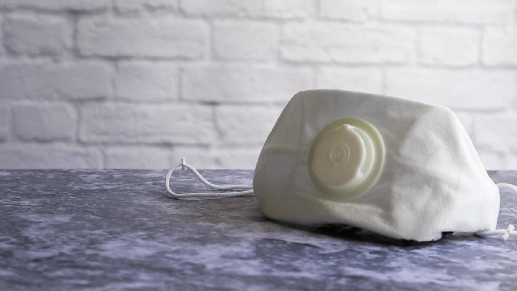 White face shield with flap on a gray table against a white brick wall with copy space. medical masks during outbreaks of viral diseases such as coronavirus or COVID-19 pandemic. photo