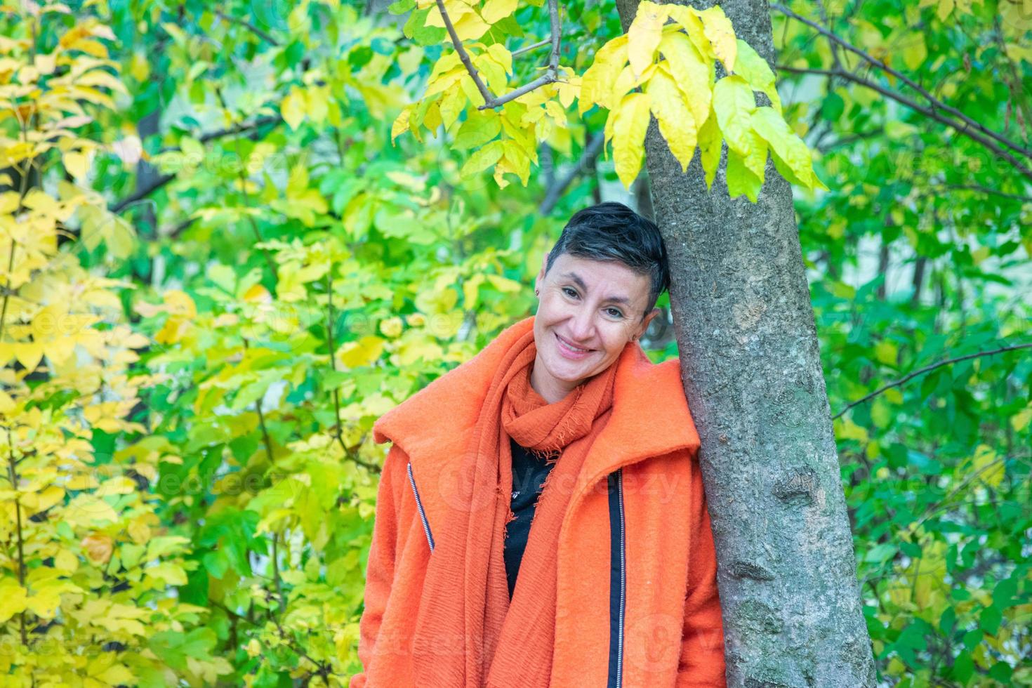 smiling middle-aged woman posing next to tree in orange coat photo