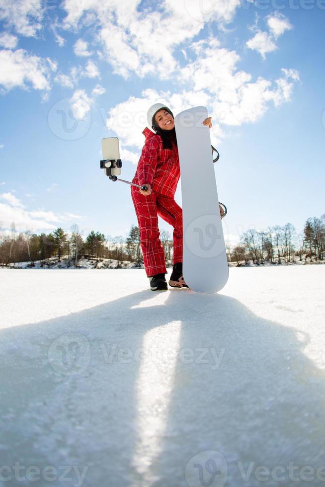 Mujer con la tabla de snowboard con smartphone en un palo para hacer selfie foto