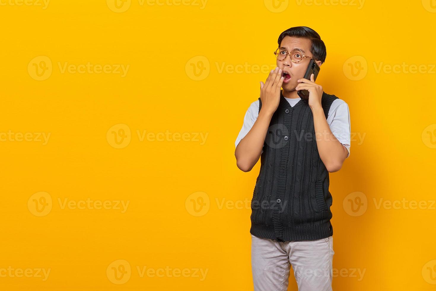 Shocked Asian young man talking on smartphone and covering mouth on yellow background photo