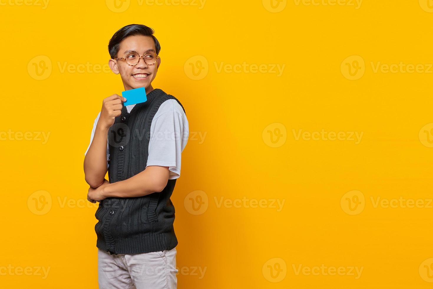Cheerful young Asian man holding credit card in hand and looking aside over yellow background photo