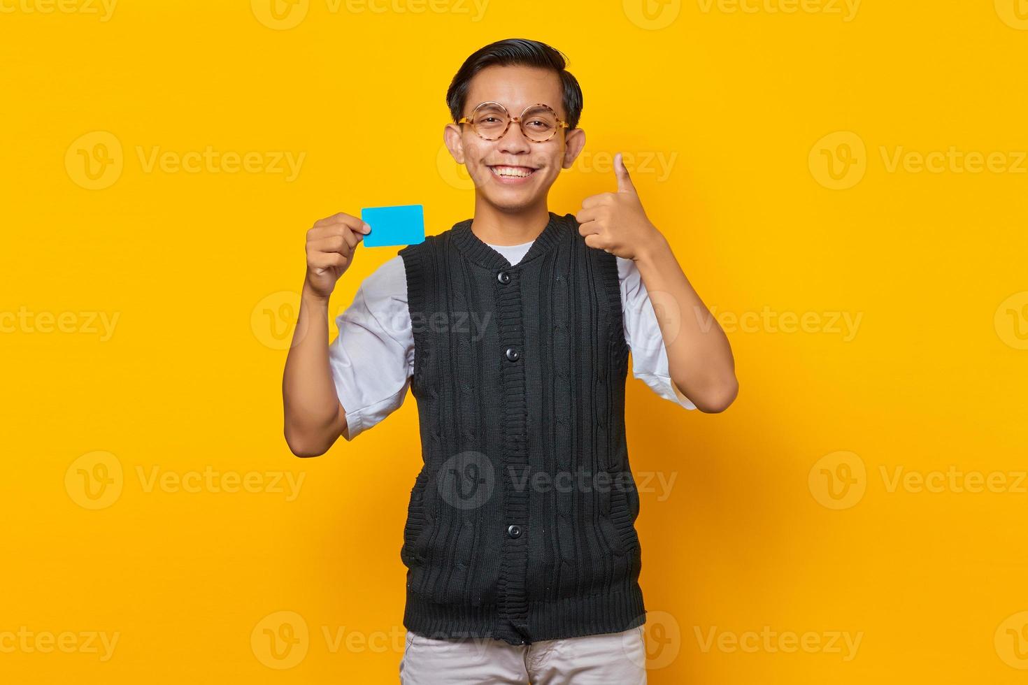 Cheerful young Asian man holding credit card and showing thumbs up gesture on yellow background photo