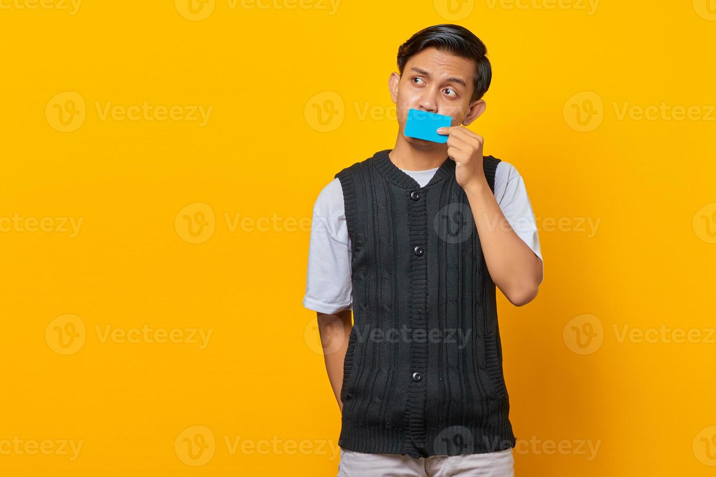 Handsome young man covering mouth with credit card and looking sideways on yellow background photo