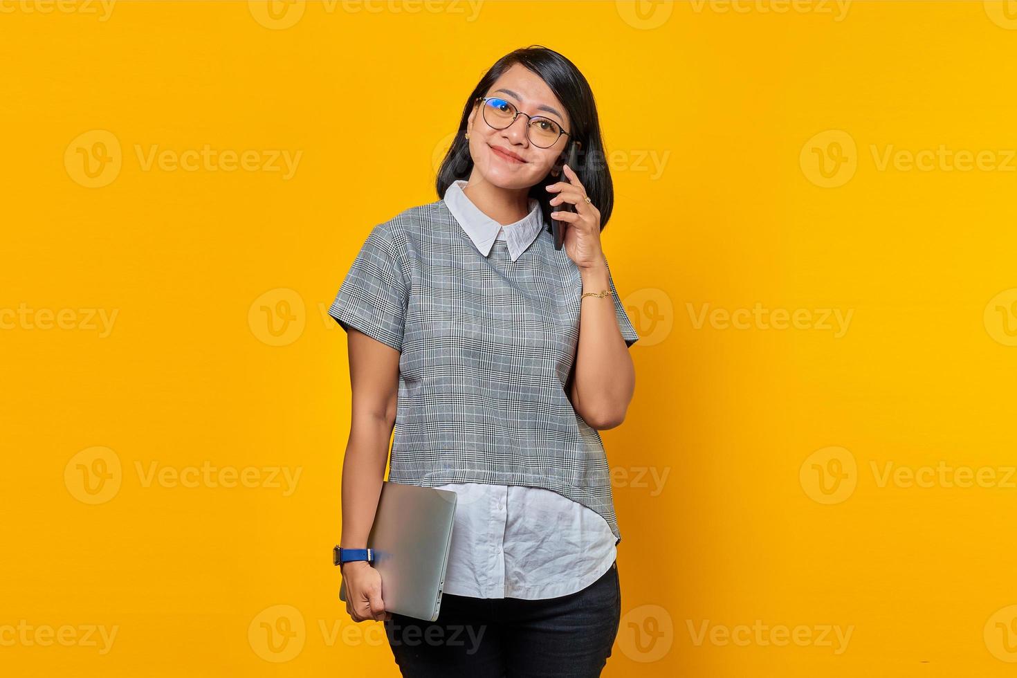 Beautiful asian woman holding laptop and talking on mobile phone over yellow background photo
