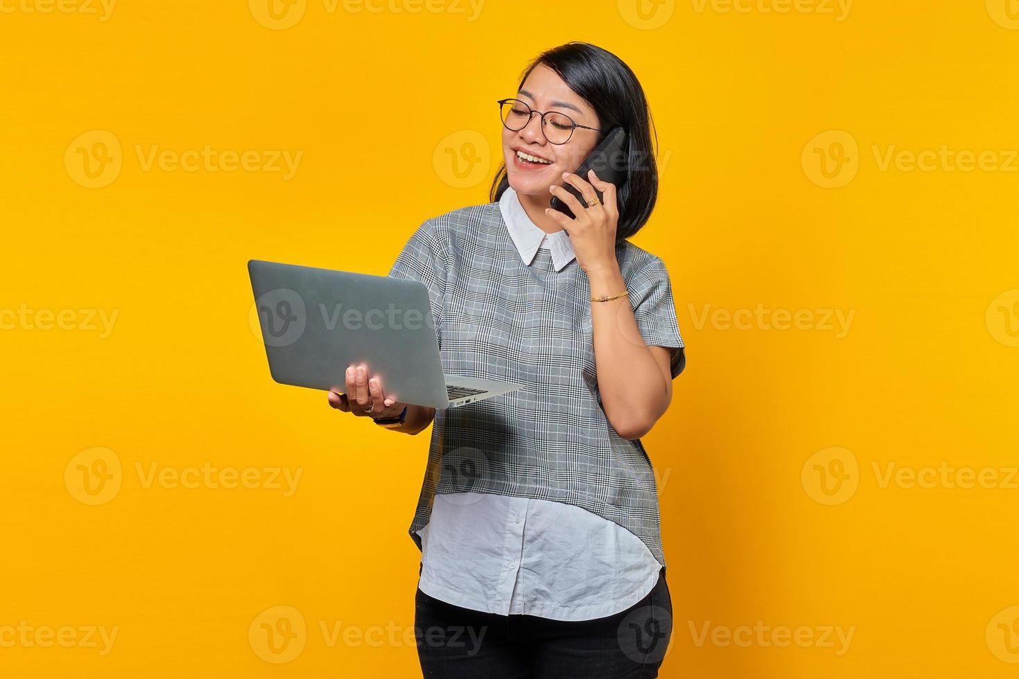 Smiling beautiful young woman talking on smartphone and looking at laptop screen on yellow background photo