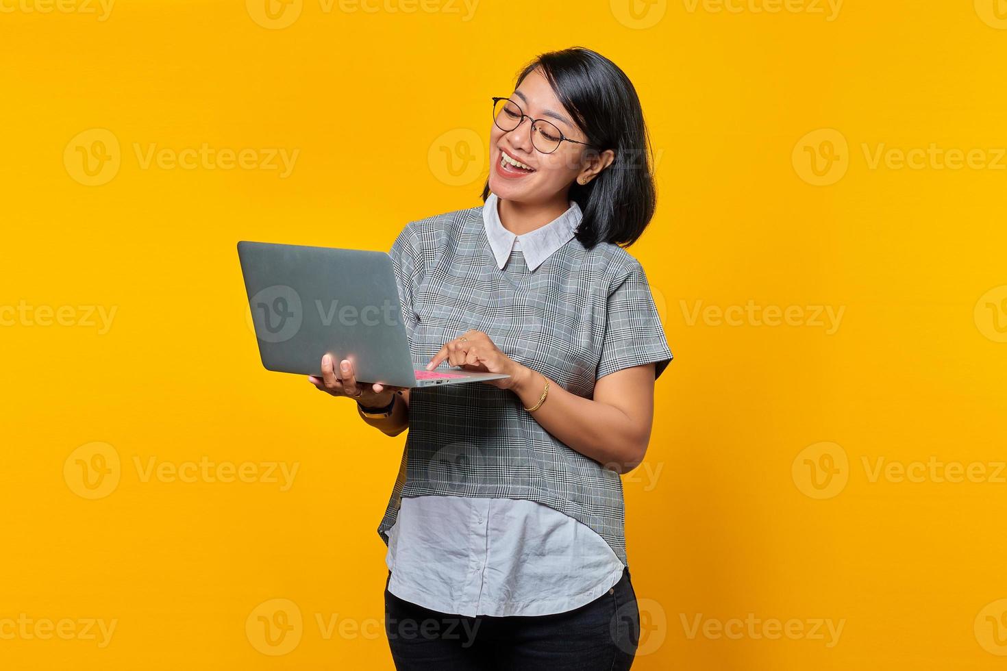 Sonriente mujer asiática con gafas leyendo el correo electrónico entrante en un portátil aislado sobre fondo amarillo foto