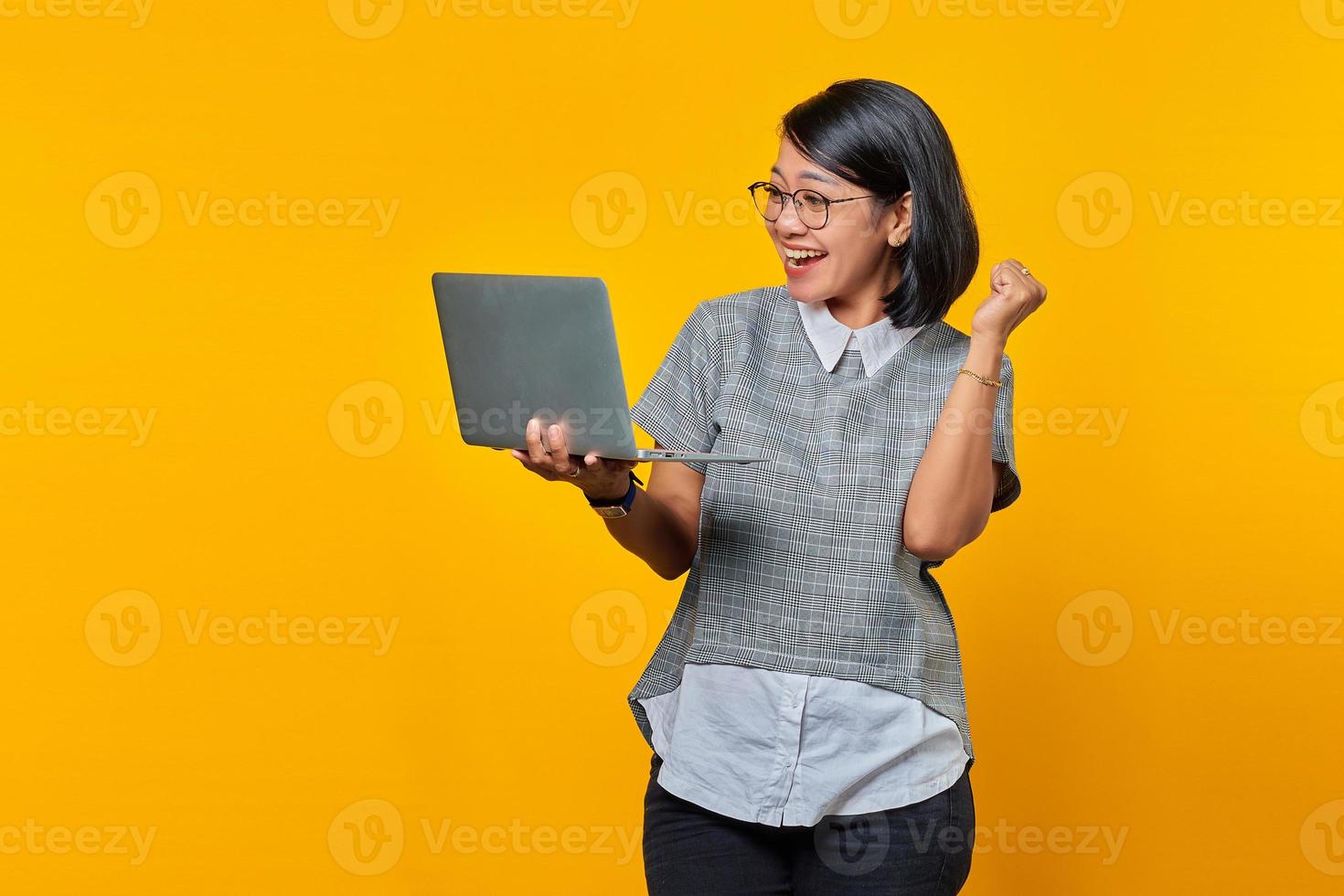 Portrait of Asian woman wearing glasses excited while opening incoming email on laptop isolated on yellow background photo