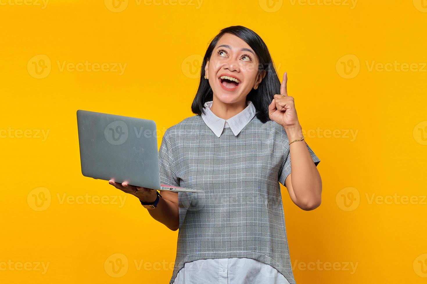 Retrato de mujer asiática emocionada sosteniendo un portátil con una gran idea y mirando sobre fondo amarillo foto