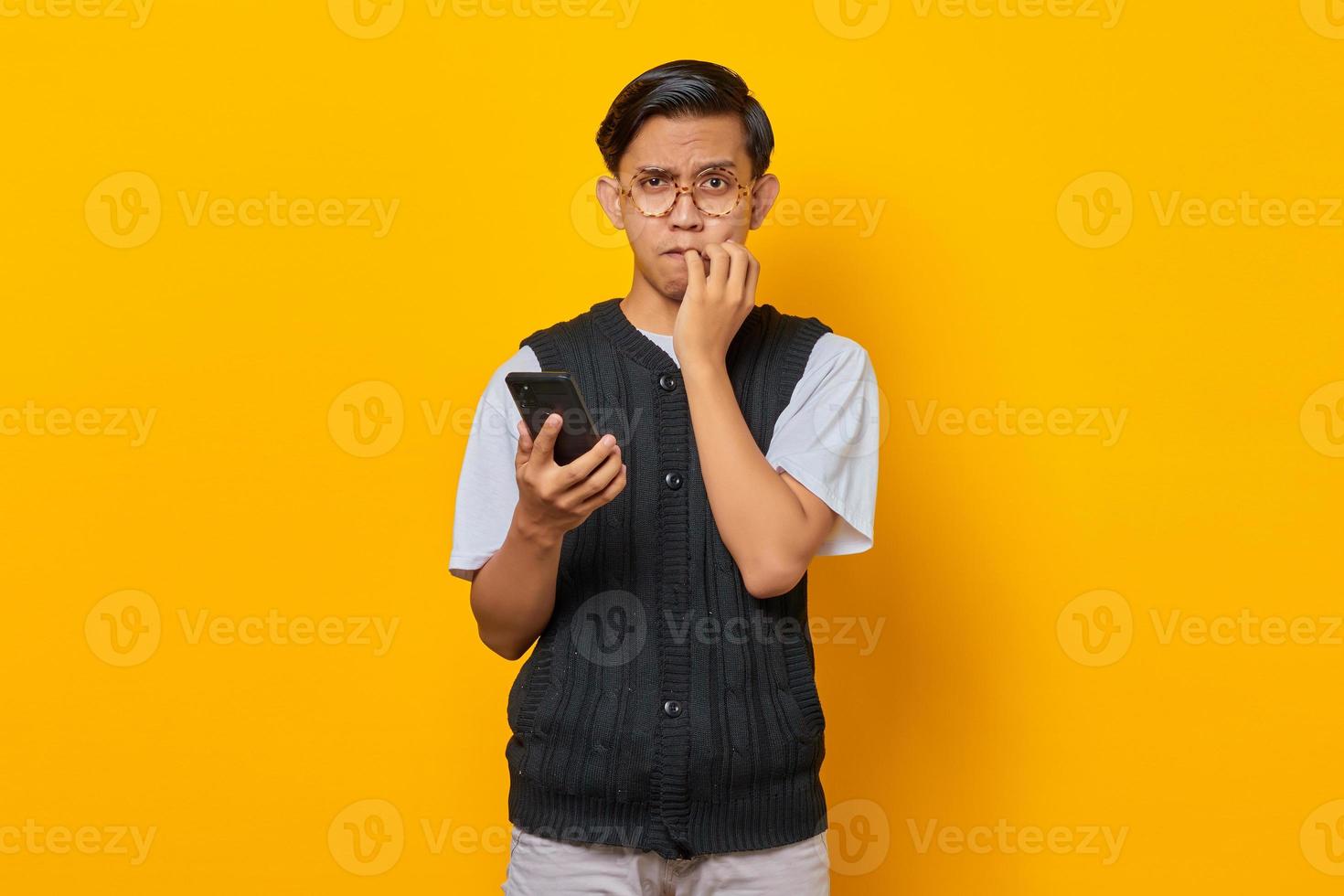 Hombre asiático joven ansioso y preocupado que sostiene el teléfono inteligente y tiene miedo de algo sobre fondo amarillo foto