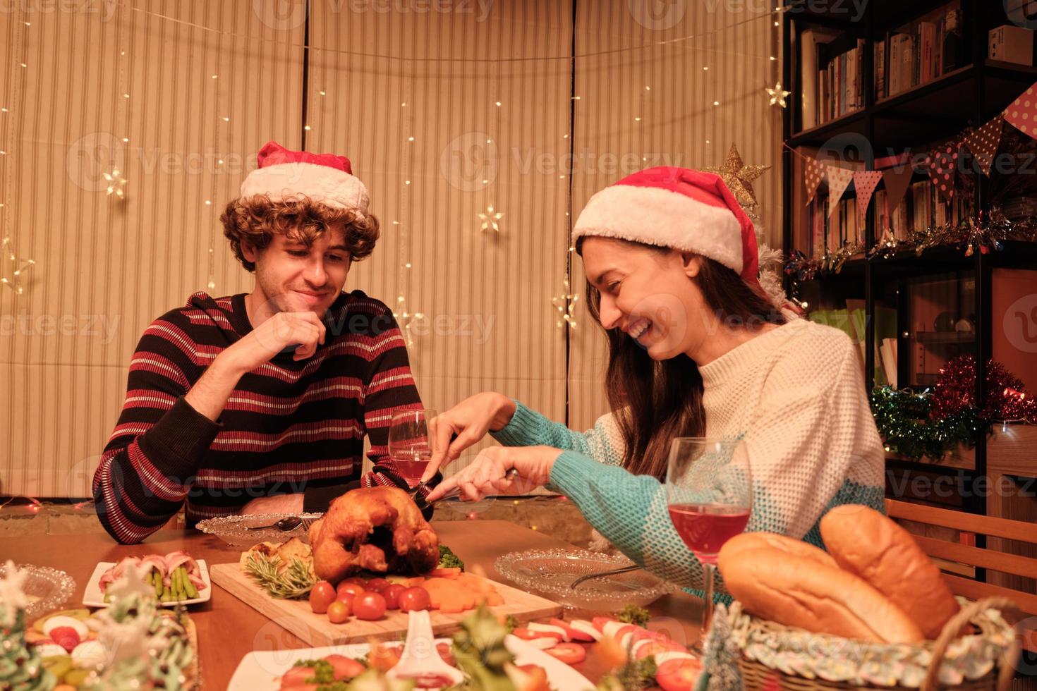 A young couple enjoys eating at table with specials foods, girlfriend cutting roasted turkey at home's dining room, decorated with ornaments, Christmas festival, and New Year celebration party. photo
