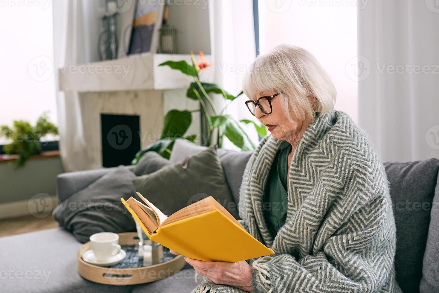 old fashioned senior woman sitting on the couch reading a book at home. Education, mature, leisure concept photo