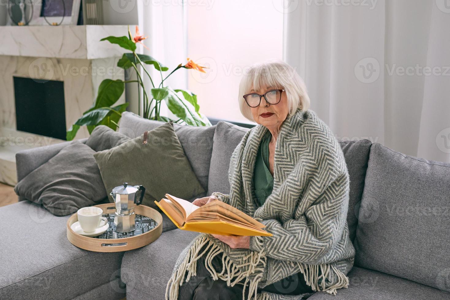 old fashioned senior woman sitting on the couch reading a book at home. Education, mature, leisure concept photo