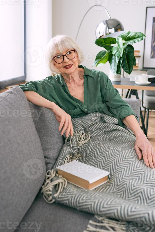 cheerful senior woman sitting on the couch reading a book at home. Education, mature, leisure concept photo