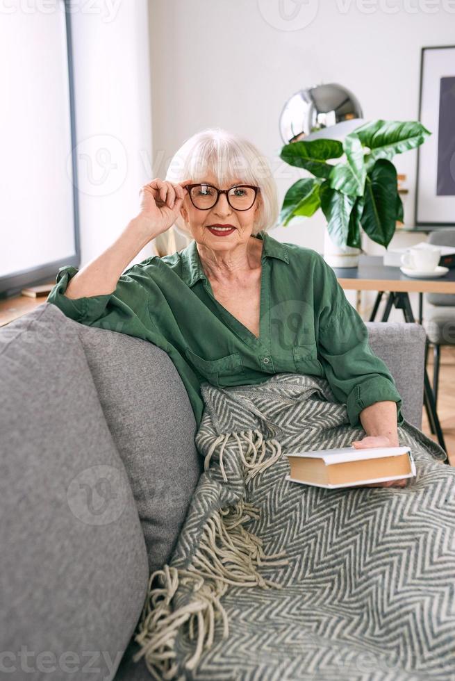 cheerful senior woman sitting on the couch reading a book at home. Education, mature, leisure concept photo