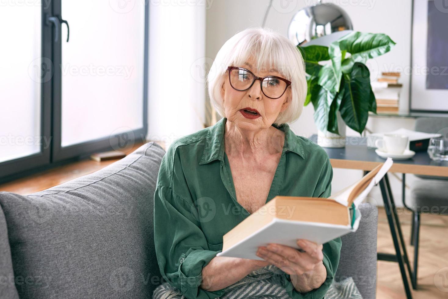 cheerful senior woman sitting on the couch reading a book at home. Education, mature, leisure concept photo