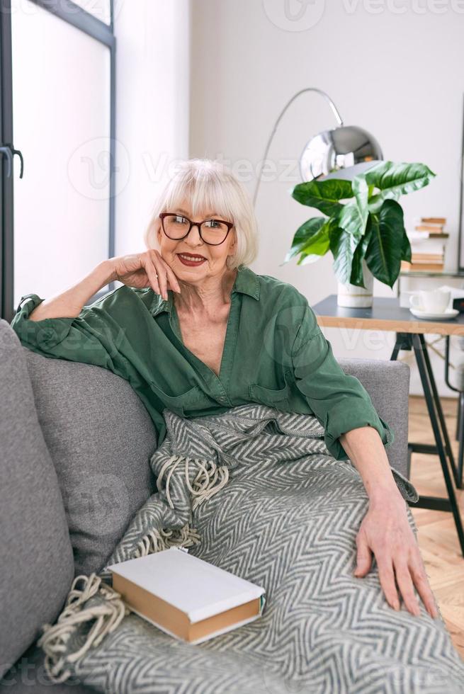 cheerful senior woman sitting on the couch reading a book at home. Education, mature, leisure concept photo