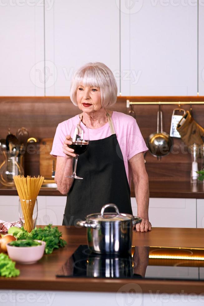 Mujer alegre senior está bebiendo vino tinto durante la cocción en la cocina moderna. comida, educación, concepto de estilo de vida foto