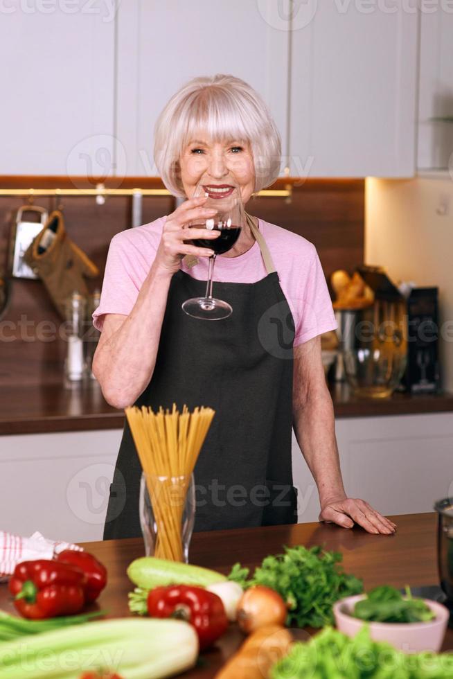 Mujer alegre senior está bebiendo vino tinto durante la cocción en la cocina moderna. comida, educación, concepto de estilo de vida foto