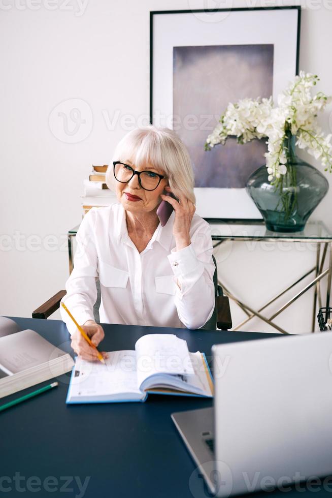 Mujer de cabello gris hermosa senior cansada en blusa blanca trabajando en equipo portátil en la oficina. trabajo, personas mayores, problemas, encontrar una solución, concepto de experiencia foto