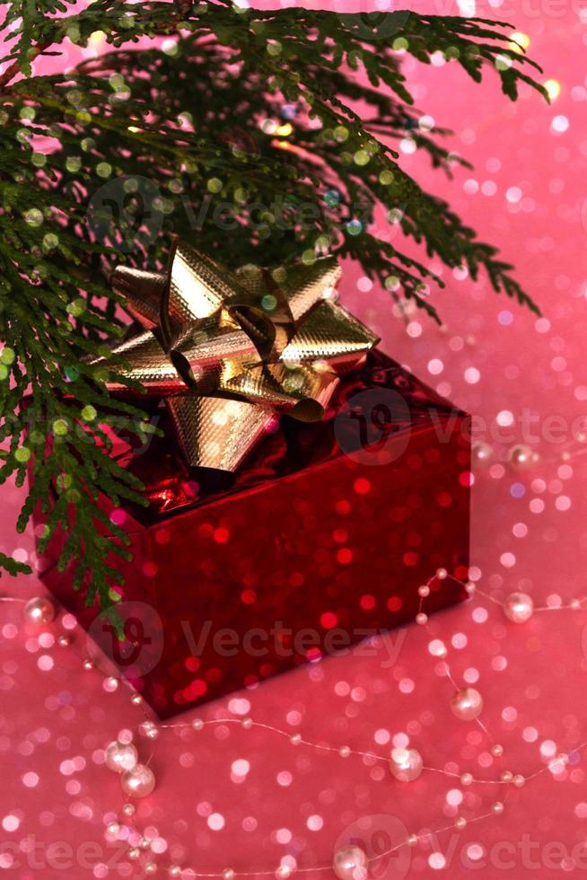 un fabuloso regalo en una caja roja con una cinta dorada debajo del árbol de navidad, sobre un fondo rosa con bokeh. año nuevo y navidad. foto