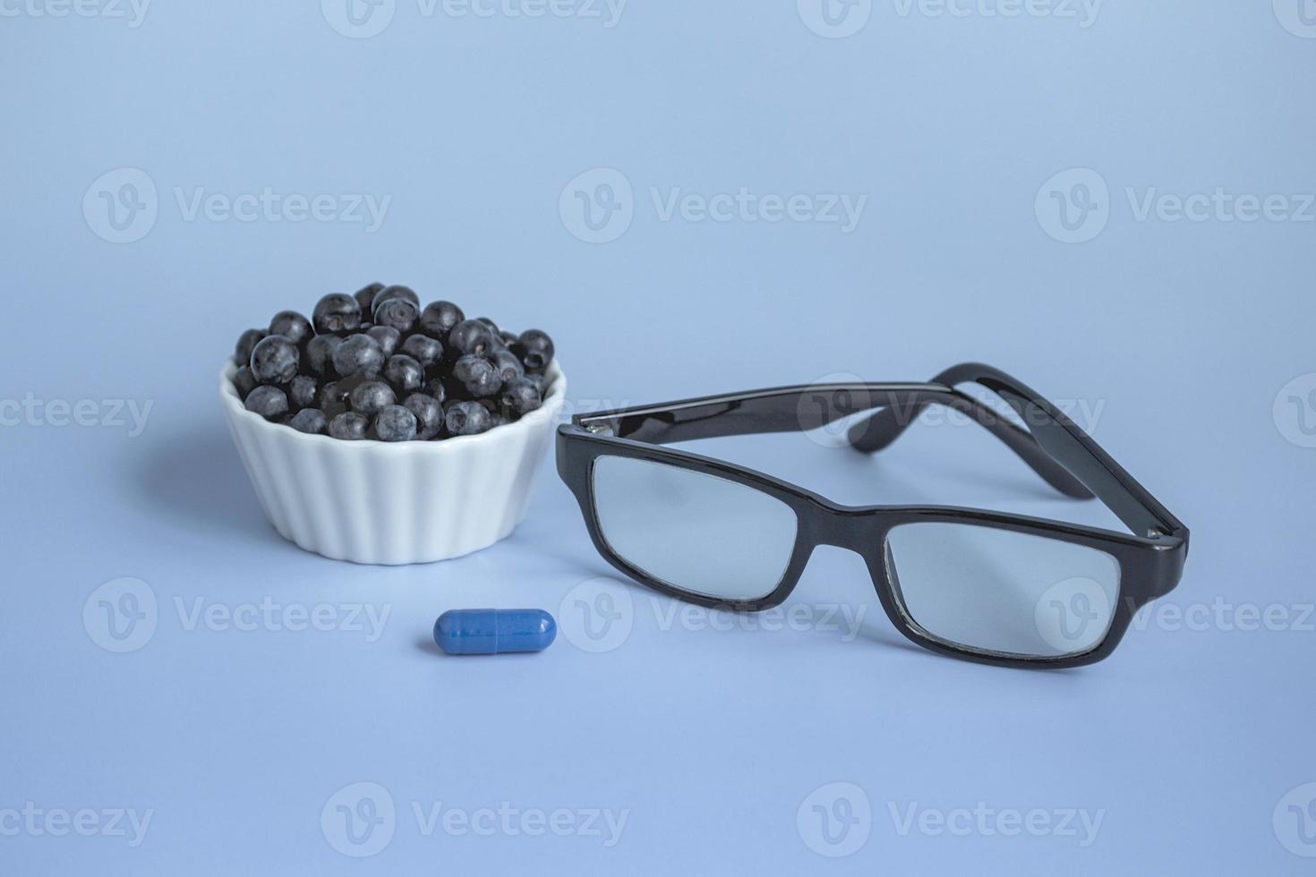 Ophthalmic concept close up. Glasses, blueberries and blueberry vitamin on a blue background. Natural products to improve and maintain vision. With copy space. photo