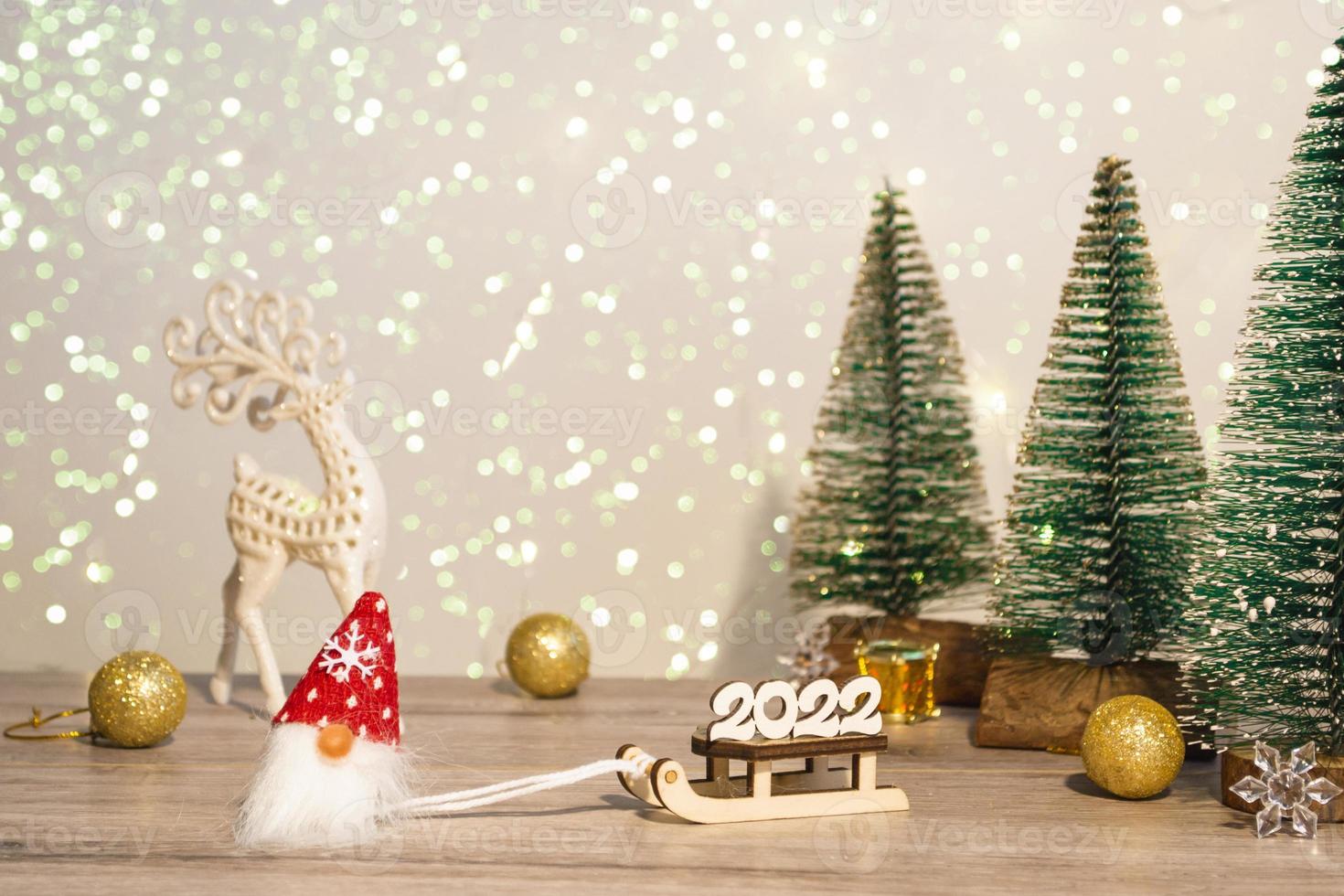 A gnome in a red hat brought the new year 2022 on a sleigh. Winter background Christmas trees in sparkles and a deer. Happy Holidays. photo
