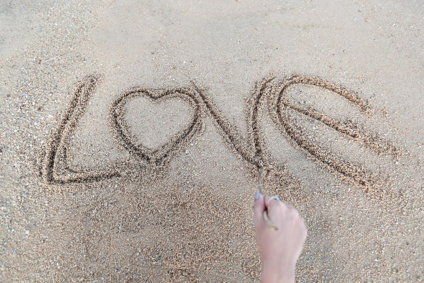 Mujer mano haciendo el símbolo del corazón y la palabra amor escrita a mano en la playa de arena foto