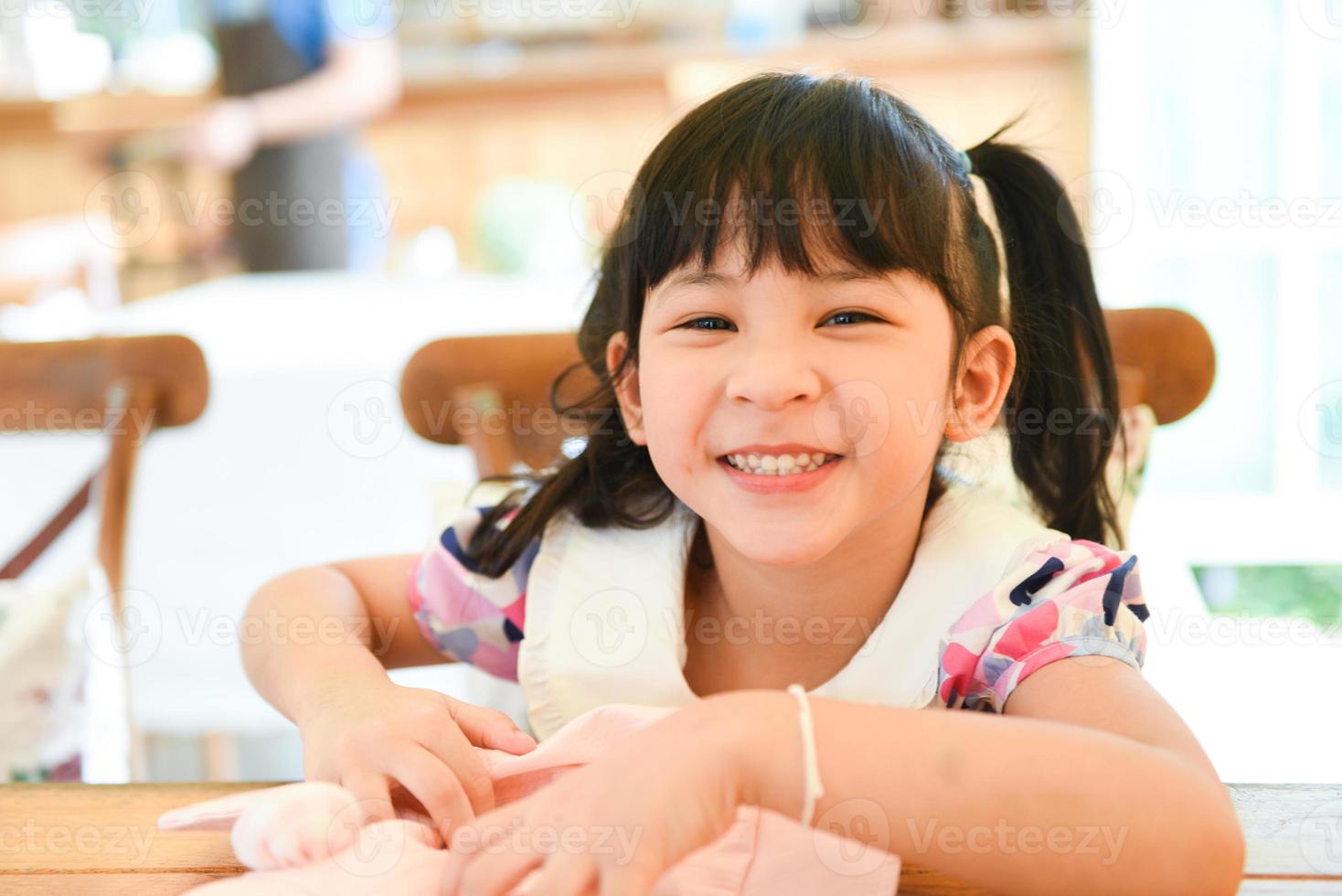 La niña asiática se divierte con una cara sonriente feliz en el café, los niños lindos de la muchacha juegan al restaurante de la mesa del comedor. foto