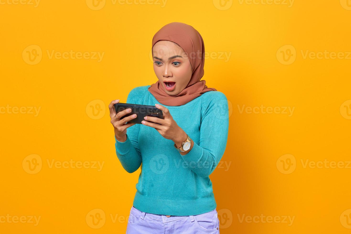 Conmocionada joven asiática jugando un videojuego en un teléfono inteligente sobre fondo amarillo foto