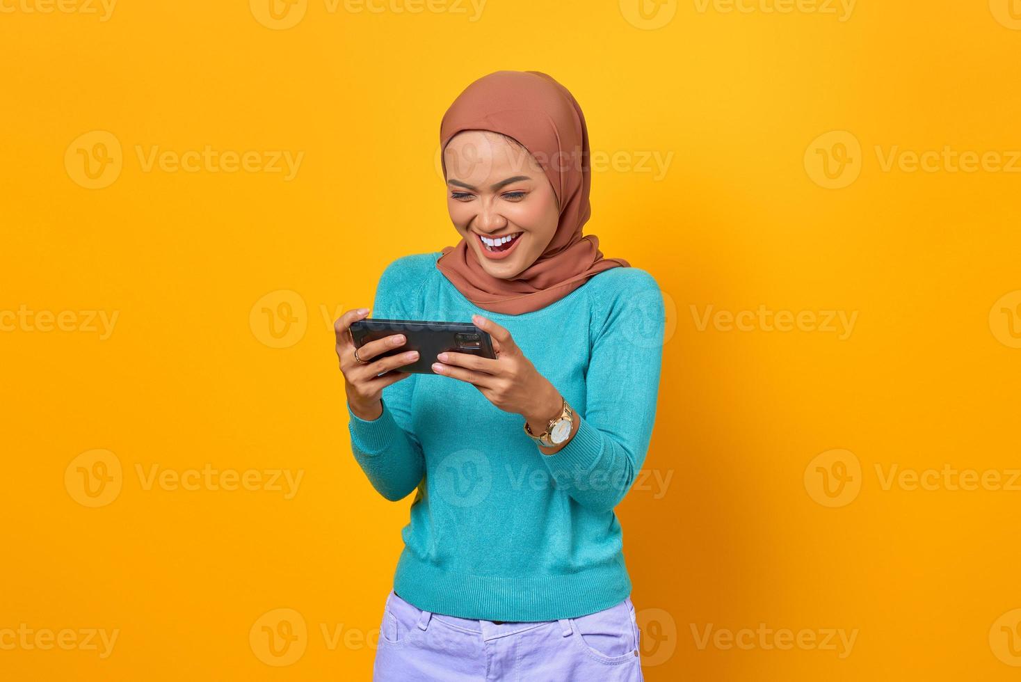 Cheerful young Asian woman playing a video game on a smartphone on yellow background photo