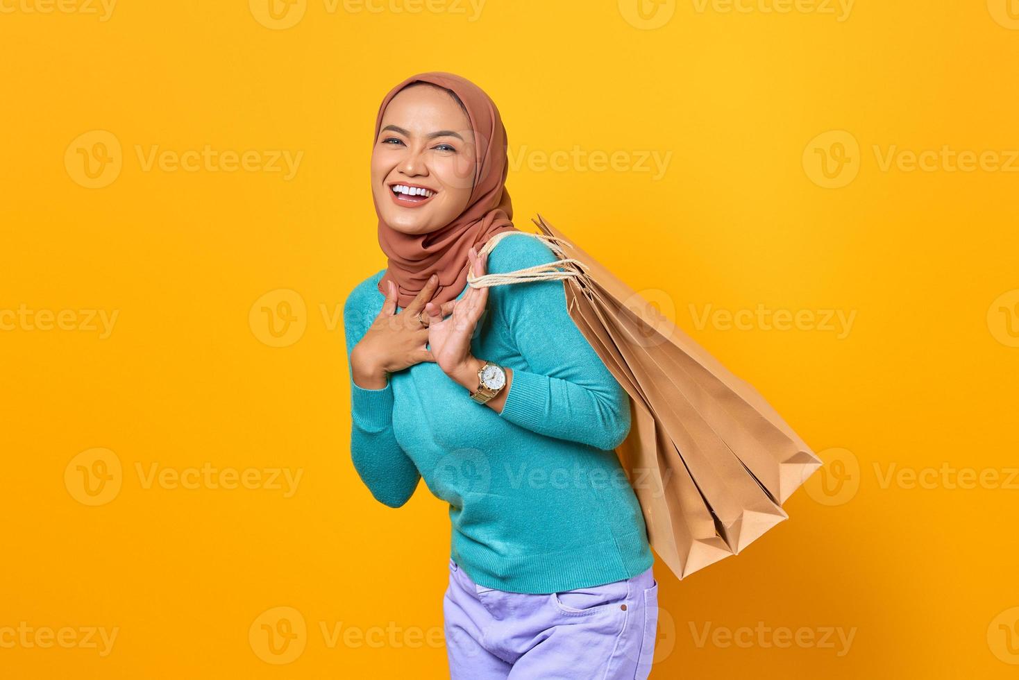 Happy young Asian woman with hand on a chest and holding shopping bags on yellow background photo