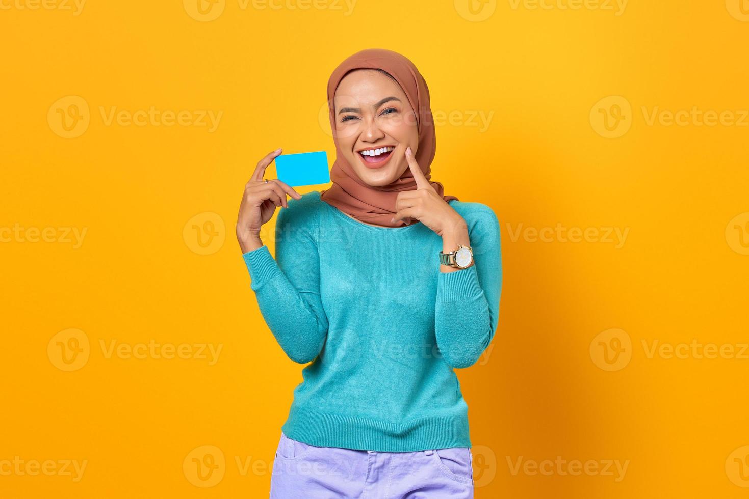 Alegre joven asiática sosteniendo una tarjeta de crédito mientras toca la mejilla sobre fondo amarillo foto