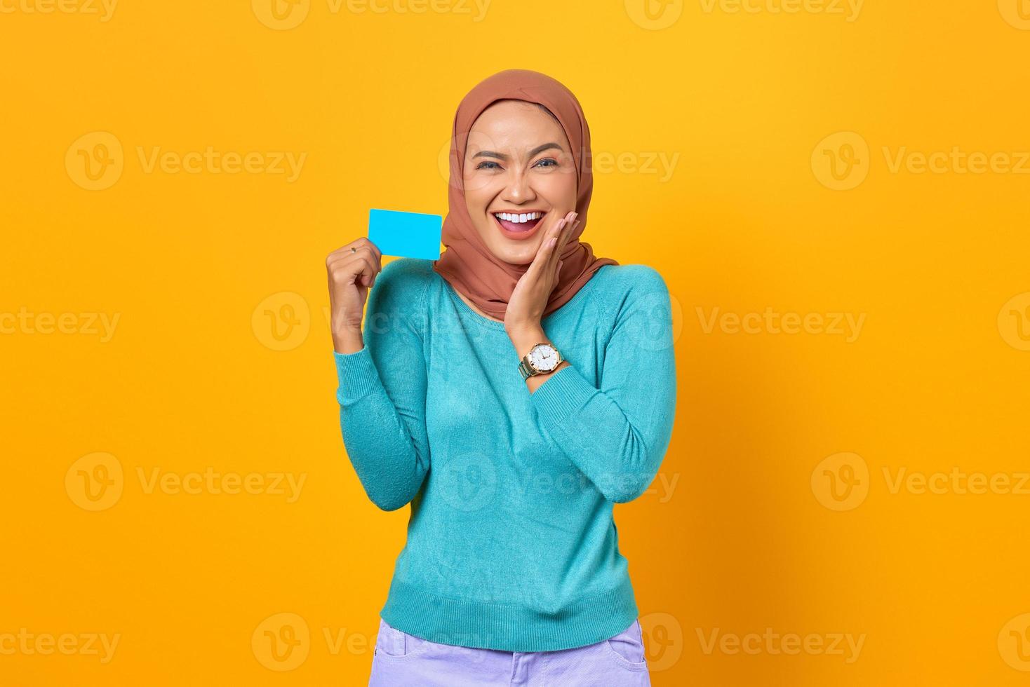Happy young Asian woman holding credit card and touch cheeks on yellow background photo