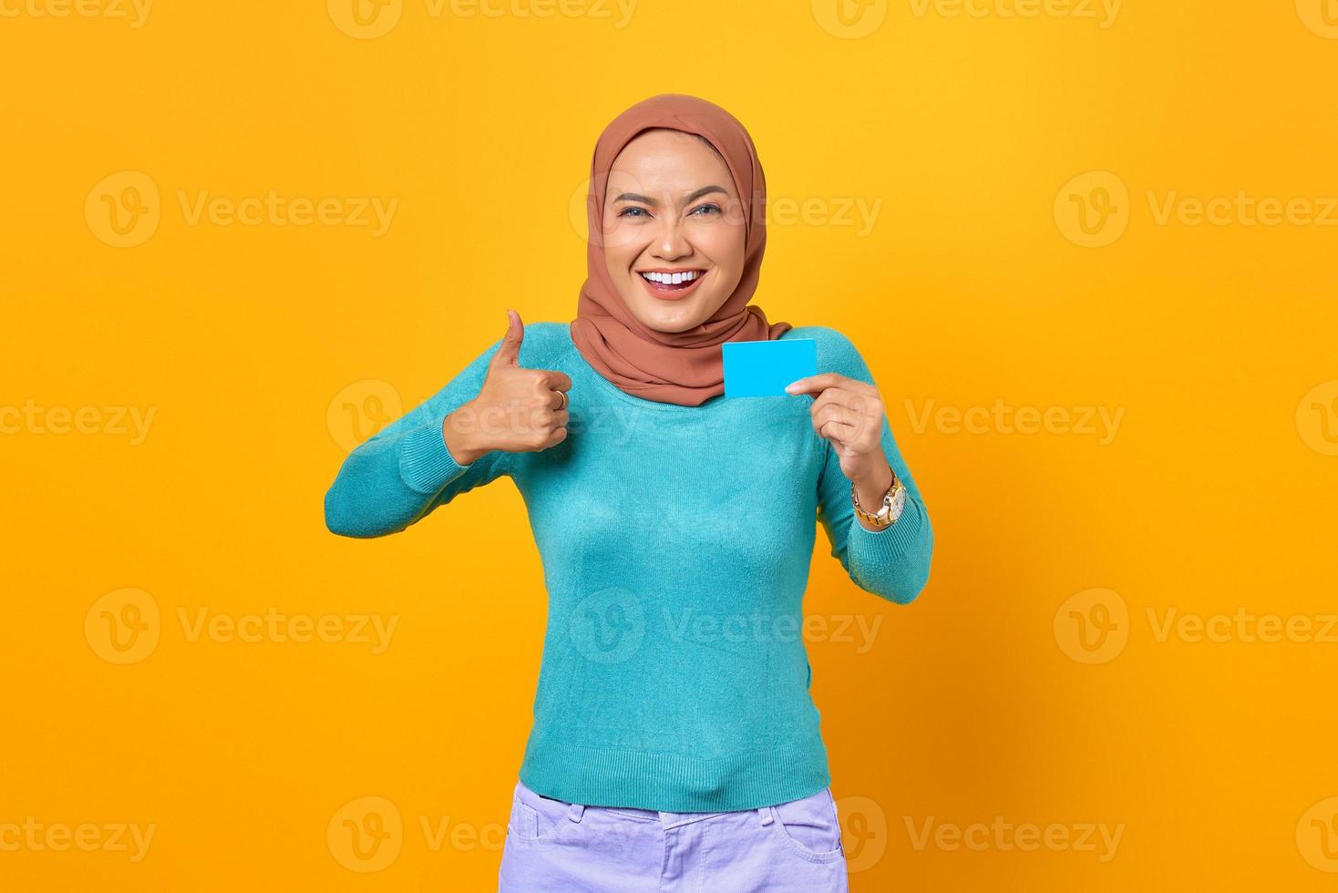 Sonriente joven mujer asiática sosteniendo una tarjeta de crédito y mostrando el pulgar hacia arriba gesto sobre fondo amarillo foto