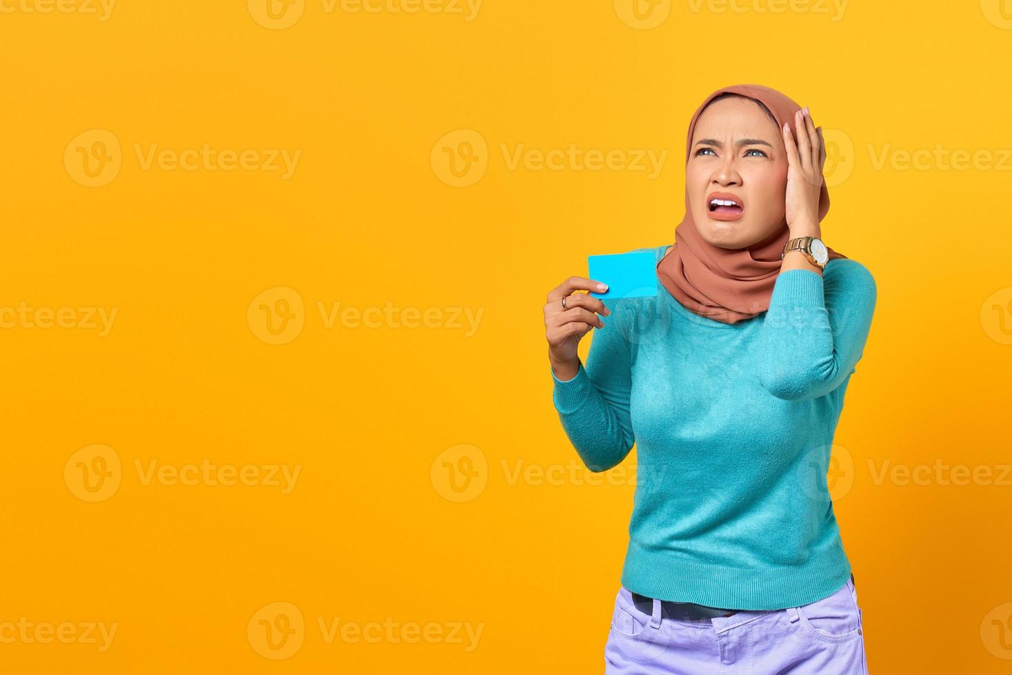 Shocked young Asian woman holding credit card and looking at copy space on yellow background photo
